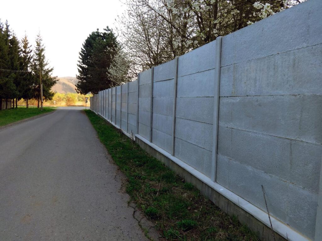 Langer, massiver Betonzaun verläuft entlang einer ländlichen Straße mit Bäumen und blühenden Sträuchern im Hintergrund