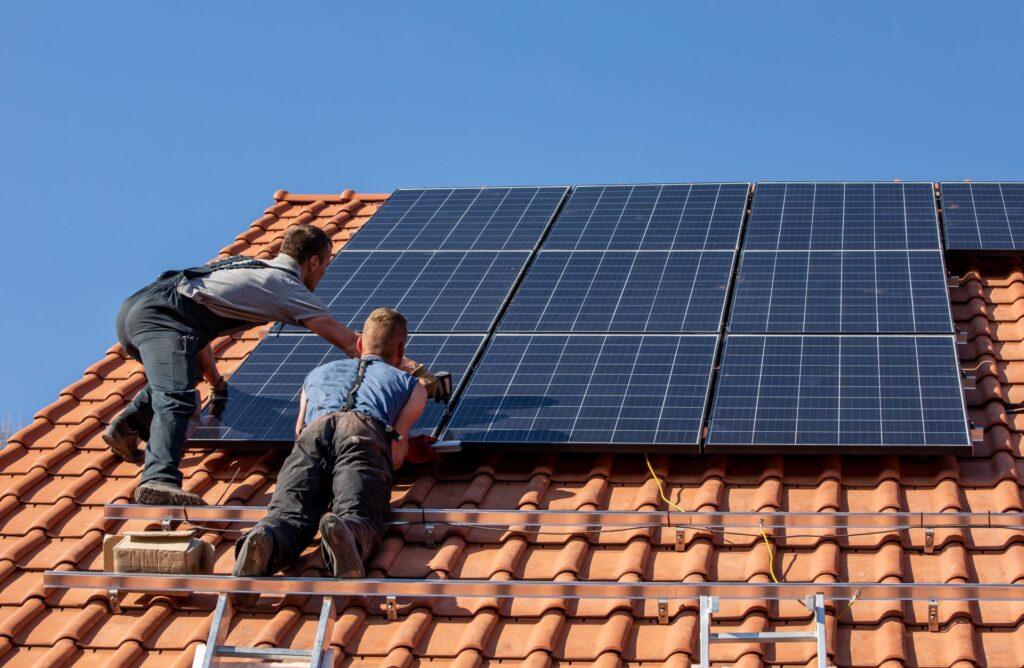 Wiederum sind zwei Arbeiter zu sehen, die Solarpaneele auf einem Ziegeldach montieren. Einer der Arbeiter hält ein Solarpanel in den Händen, während der andere es für die Installation vorbereitet. Auf dem Dach sind bereits einige Paneele installiert, und die Szene spielt sich an einem sonnigen Tag ab.