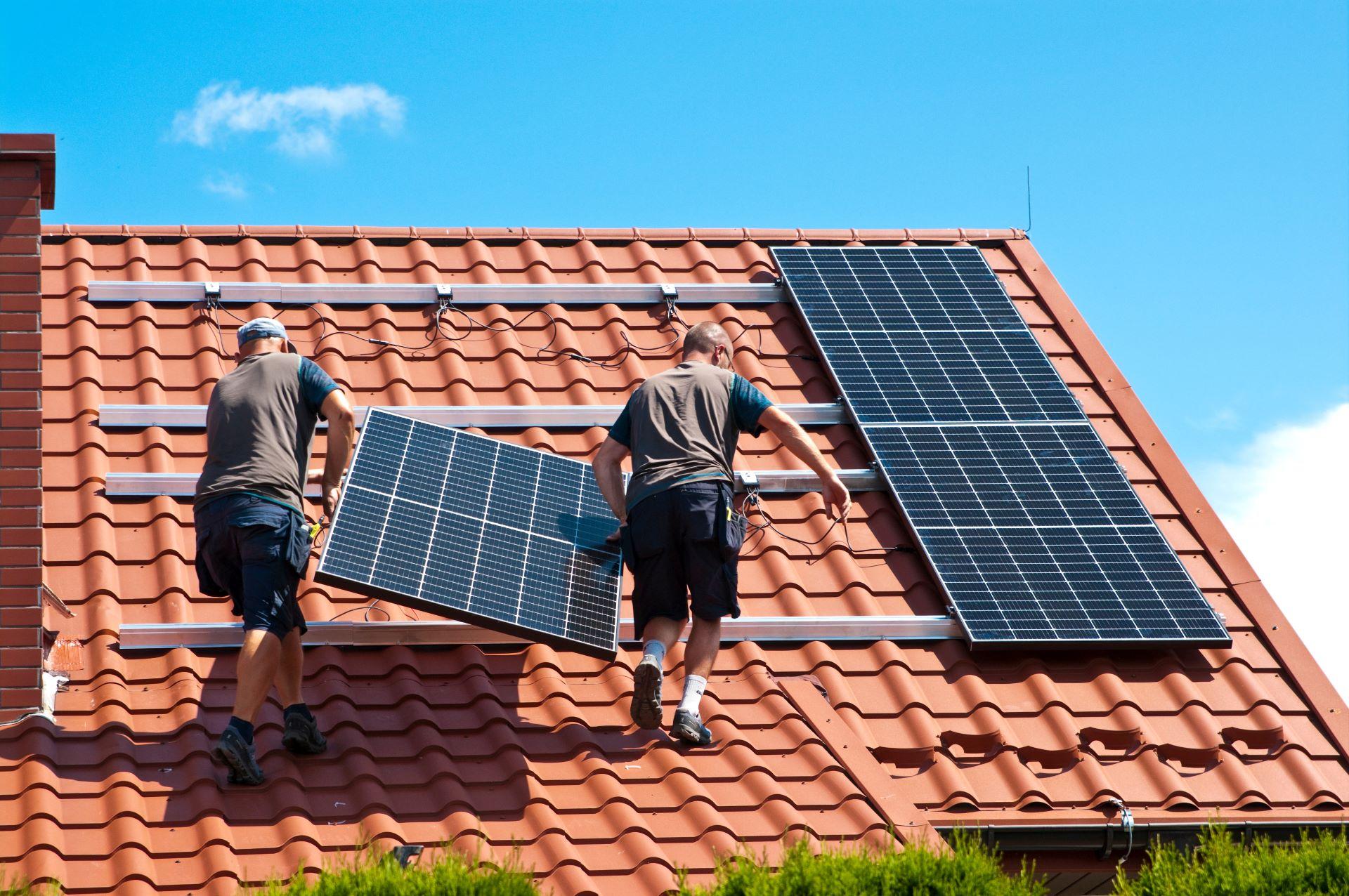 Zwei Arbeiter montieren Solarmodule auf einem Ziegeldach. Sie benutzen Leitern und Sicherheitsausrüstung, um die Solarpaneele auf dem Dach zu befestigen. Das Bild zeigt die Installation von Solarpanelen, um das Haus mit erneuerbarer Energie zu versorgen.