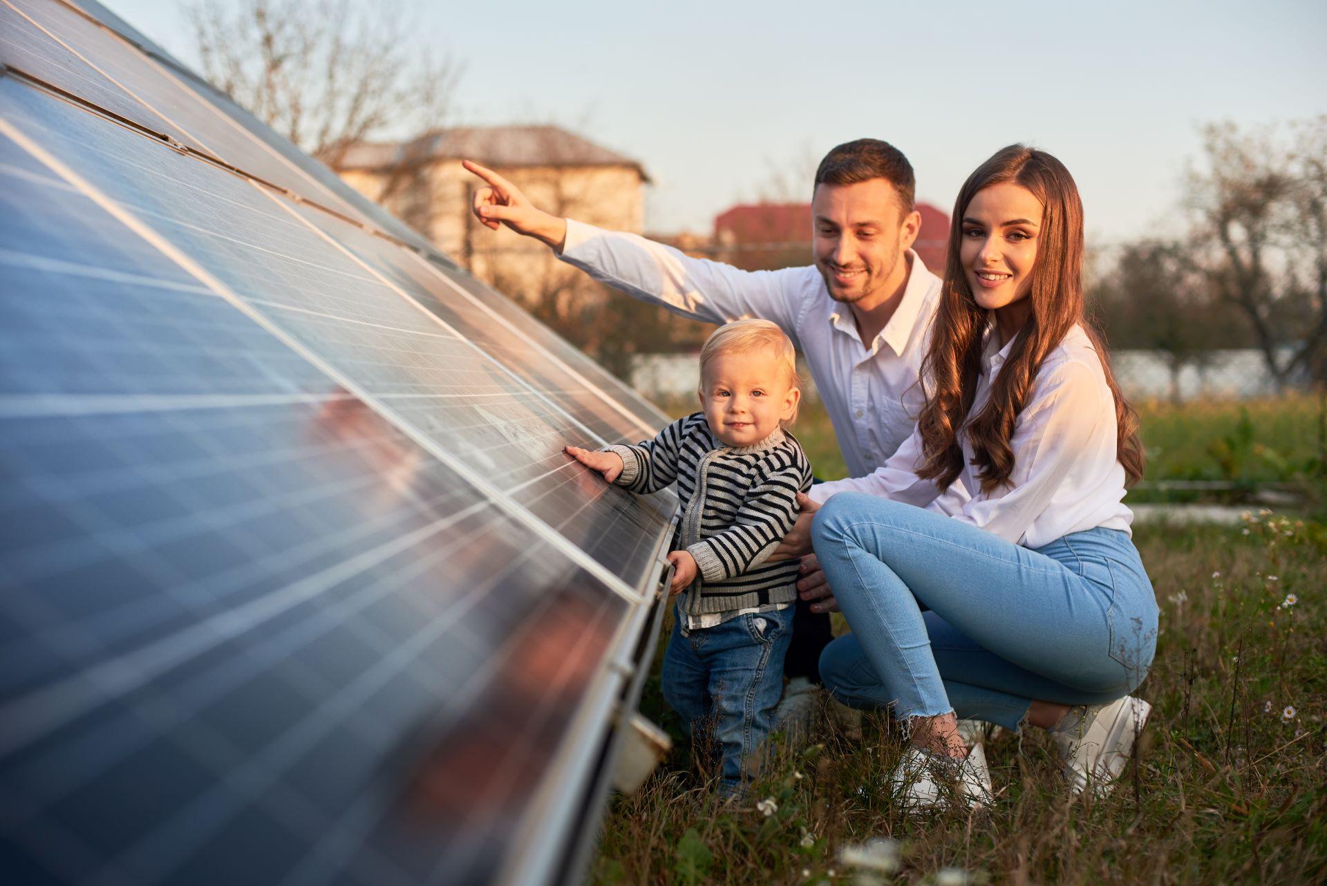 Eine Familie bestehend aus Mutter, Vater und einem kleinen Kind steht vor Solarmodulen, die auf dem Boden installiert sind. Das Kind berührt eines der Paneele, während der Vater auf etwas in der Ferne zeigt und die Mutter lächelt. Es handelt sich wahrscheinlich um eine familiäre Umgebung, möglicherweise ihr Zuhause, und symbolisiert das Engagement der Familie für erneuerbare Energien.