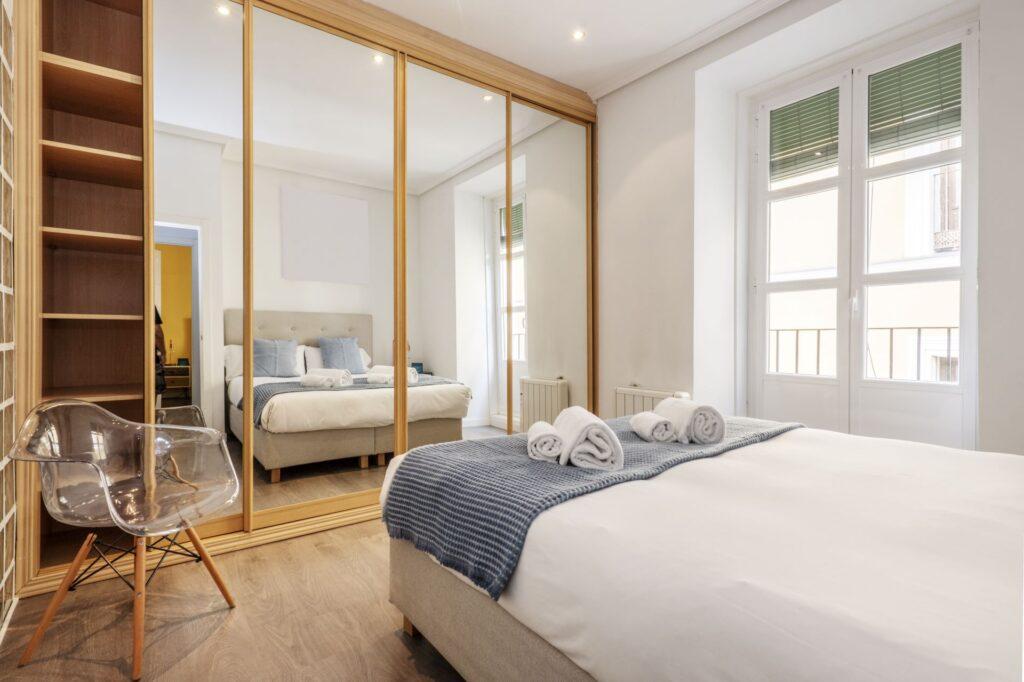 Helles Schlafzimmer mit großem Kleiderschrank mit Spiegelfront und Schiebetüren, Doppelbett mit gefalteten Handtüchern, Holzfußboden und Tageslicht.
